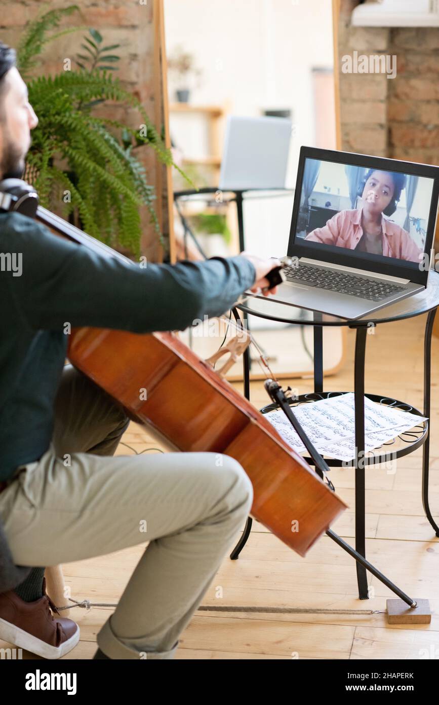 Adorable écolier africain sur l'écran d'un ordinateur portable regardant un professeur de musique jouant du violoncelle tout en étant assis à la maison Banque D'Images