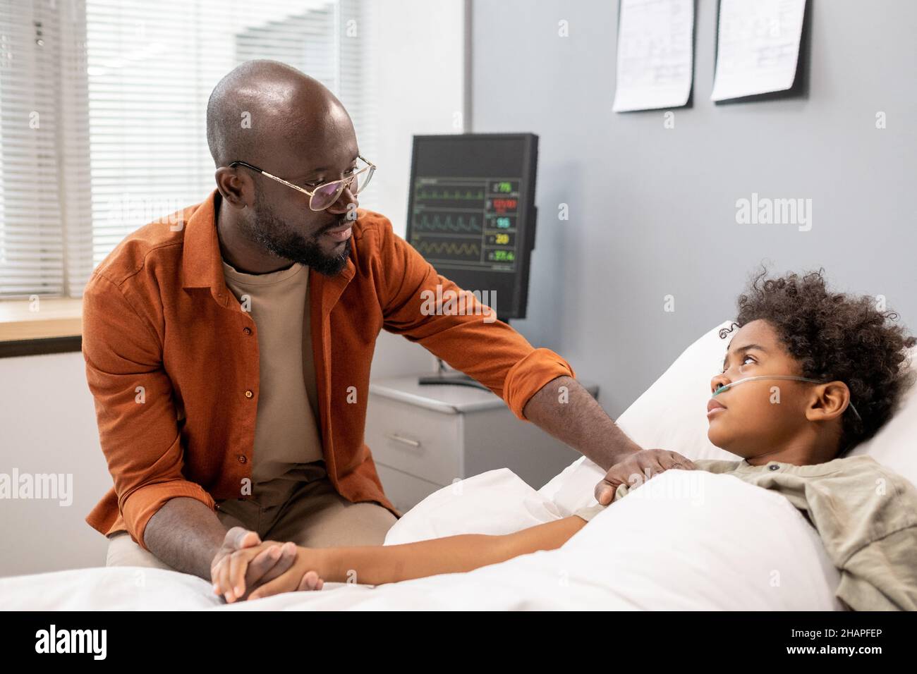 Jeune père africain soutenant son fils pendant la maladie, il lui parlant alors qu'il est allongé au lit à l'hôpital Banque D'Images