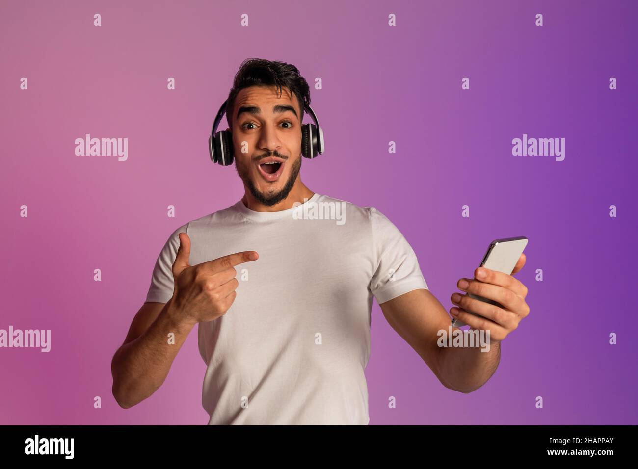 Surpris jeune arabe gars dans le casque pointant vers smartphone, choqué par la vente en ligne dans la lumière de néon Banque D'Images