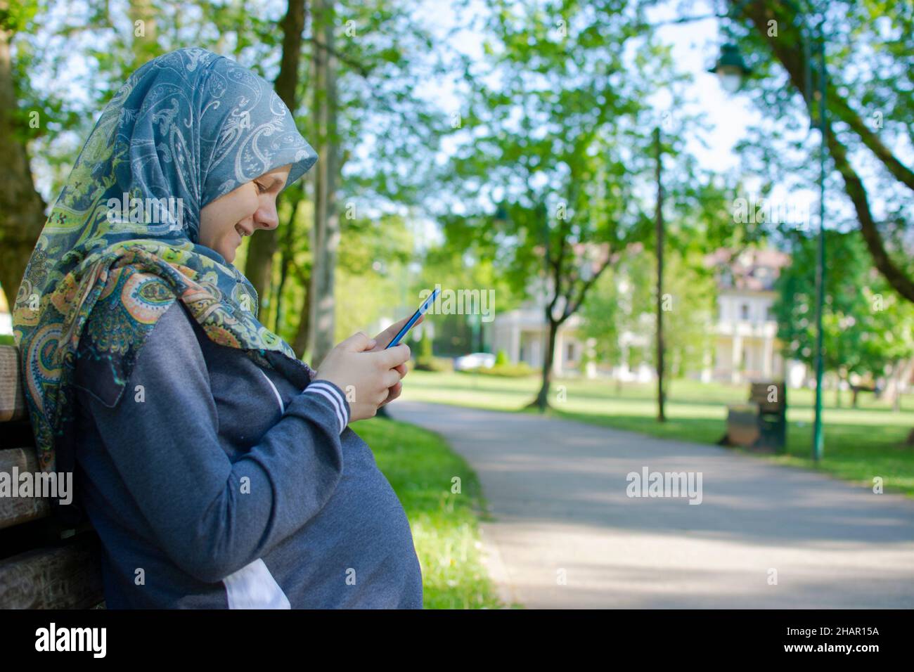 Vue de profil de la femme enceinte musulmane souriant tout en utilisant un téléphone portable et assis seul dans le parc Banque D'Images