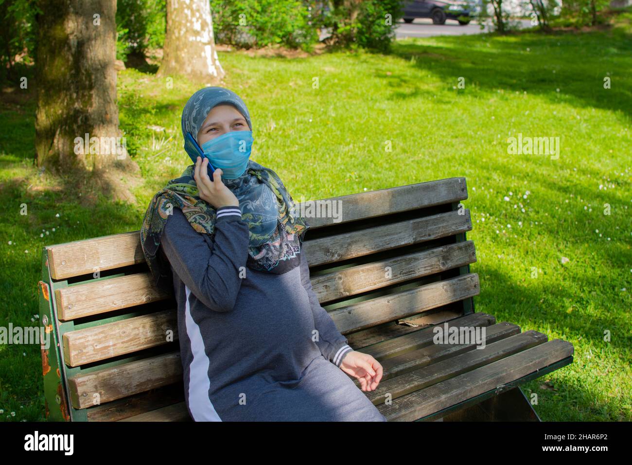 Jeune femme enceinte musulmane portant un masque facial parlant au téléphone pendant qu'elle est assise dans le parc Banque D'Images