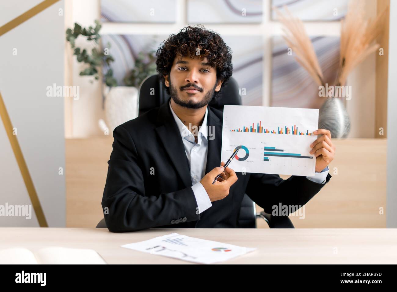 Smart réussi curly-hful Indian Business leader, mentor financier, directeur d'entreprise parler à un collègue ou client lors d'une conférence téléphonique, réunion virtuelle, brainstorming, tient des documents, regarder la caméra Banque D'Images