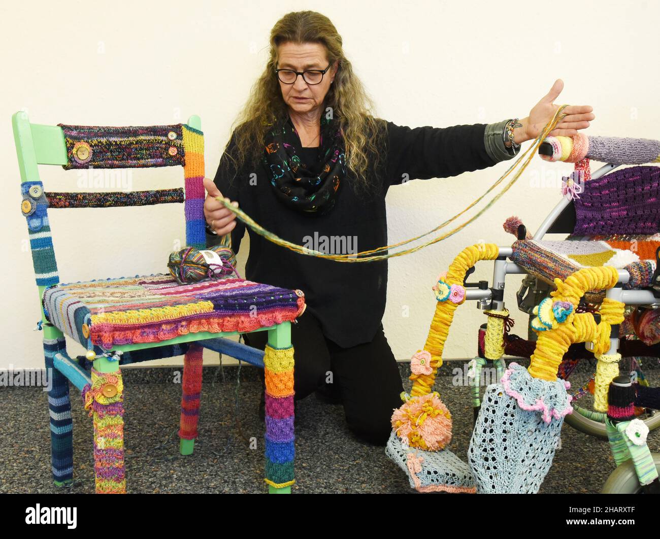 14 décembre 2021, Saxe, Torgau: La travailleuse sociale Ina Baumann-Kahlo tient un "fil de patience" dans son bureau à côté d'un fauteuil roulant d'enfant tricoté avec de la laine de couleur (r) et une chaise également tricotée dans ses mains.Pendant la pandémie de Corona, Baumann-Kahlo a passé de nombreuses heures à tricoter dans le fauteuil roulant pour remercier les nombreux aides de la crise qui n'étaient pas aussi présentes dans l'œil public, a-t-elle dit.Les gens voient le fauteuil roulant, comme elle le fait, comme un symbole de besoin d'aide.Elle voit la chaise comme une « chaise d'aide ».Avec elle, elle veut vous remercier aux nombreux aides qui aident tranquillement les autres, même dans Banque D'Images