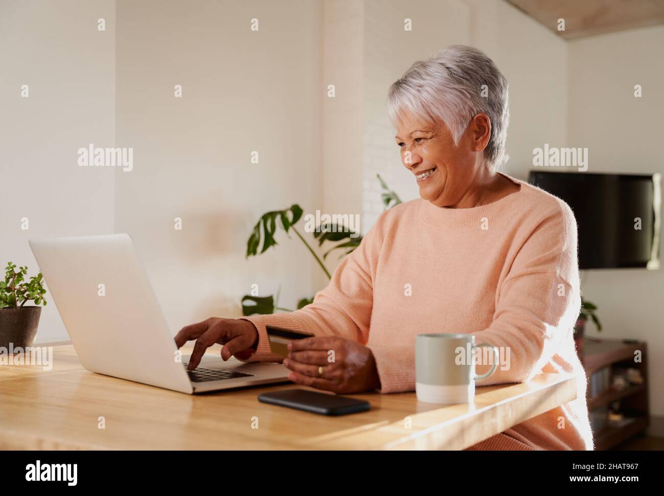 Une femme âgée de plusieurs ethnies se fait un plaisir de payer en ligne sur un ordinateur portable, assise au comptoir de cuisine, banque en ligne. Banque D'Images