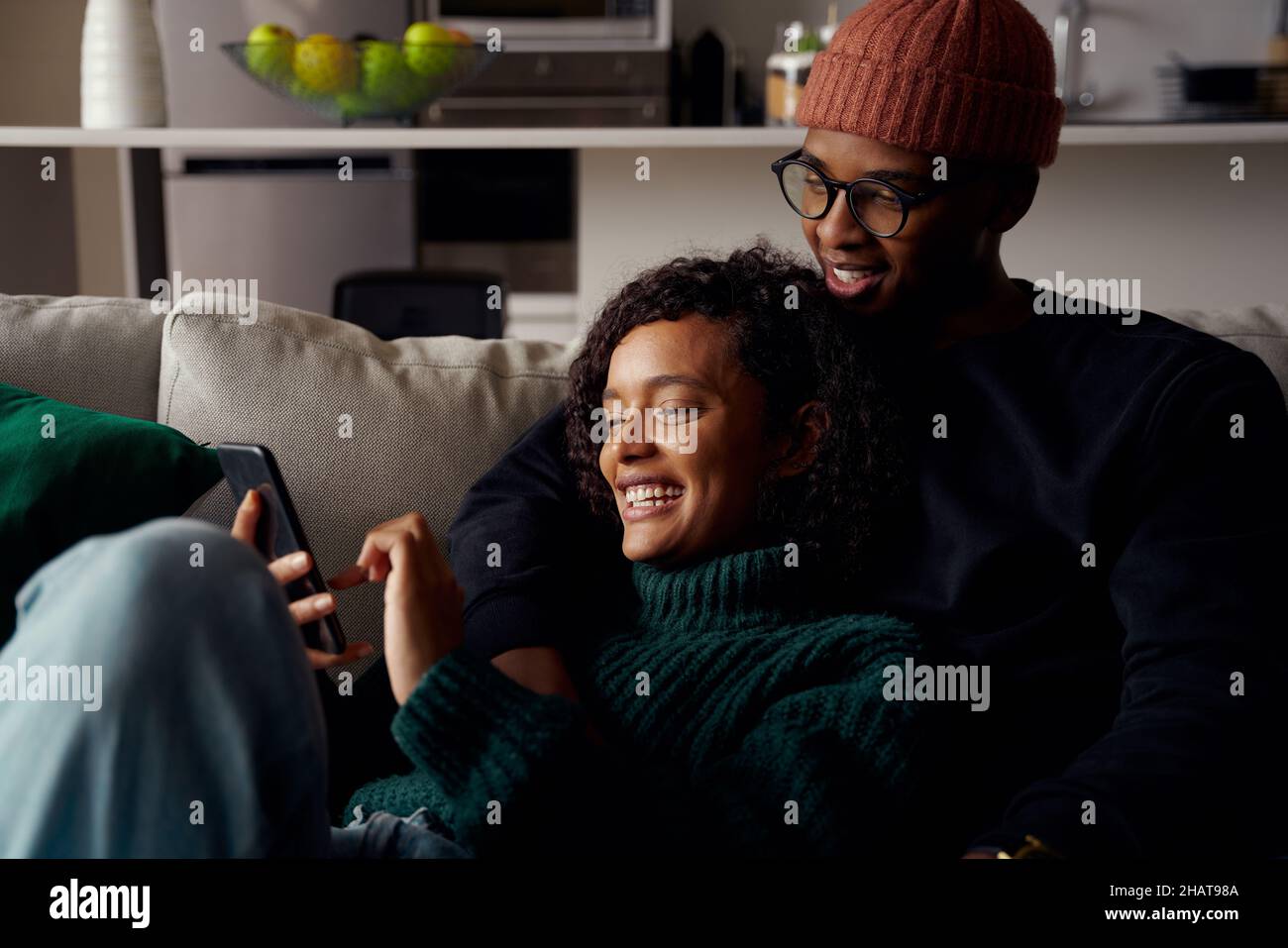 Un couple multiethnique se moque et riant du texte sur le téléphone tout en étant sur le canapé dans un appartement moderne Banque D'Images