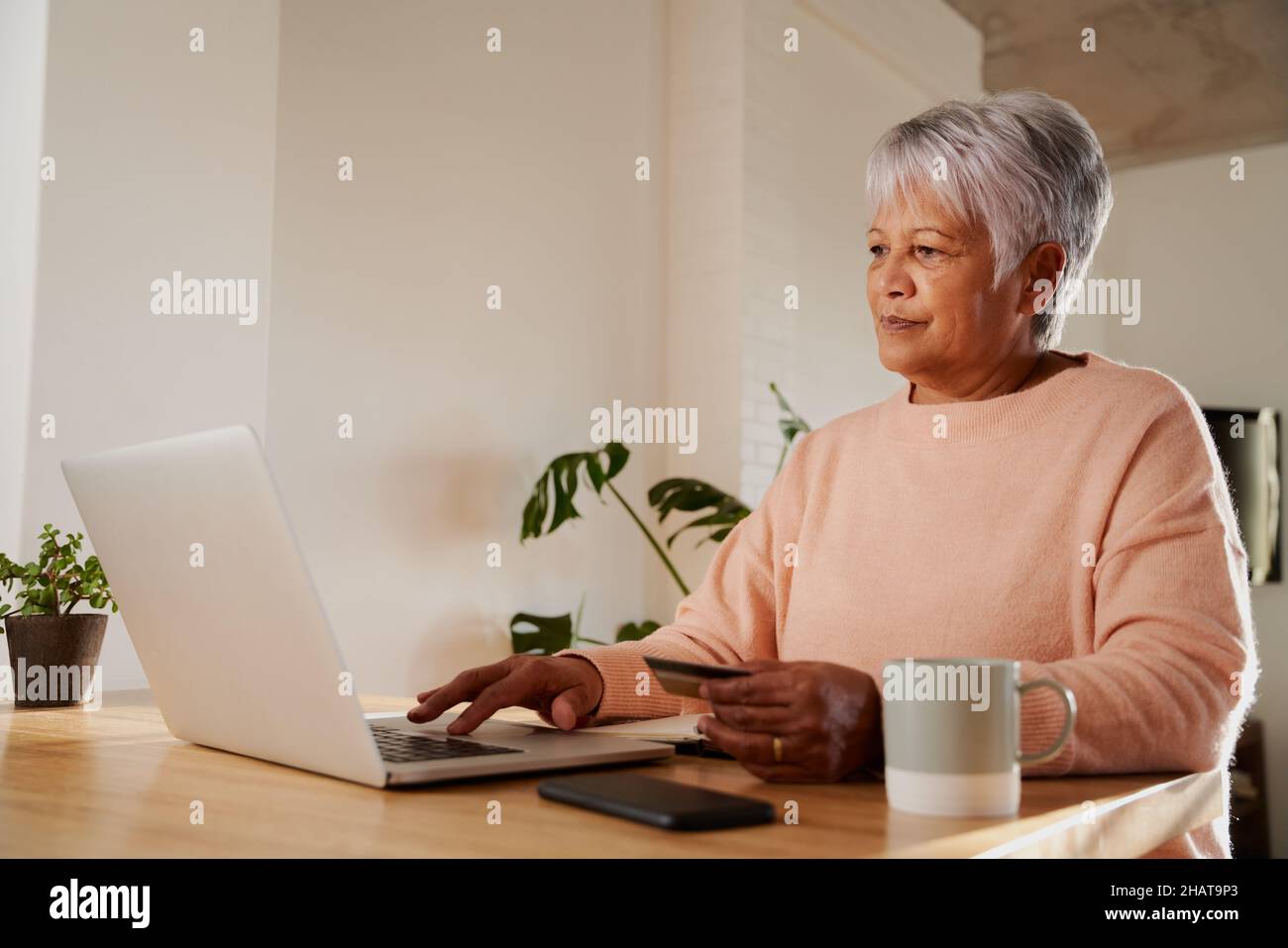 Femme âgée, multiethnique, tapant dans les détails bancaires sur un ordinateur portable, assise au comptoir de cuisine moderne. Banque D'Images