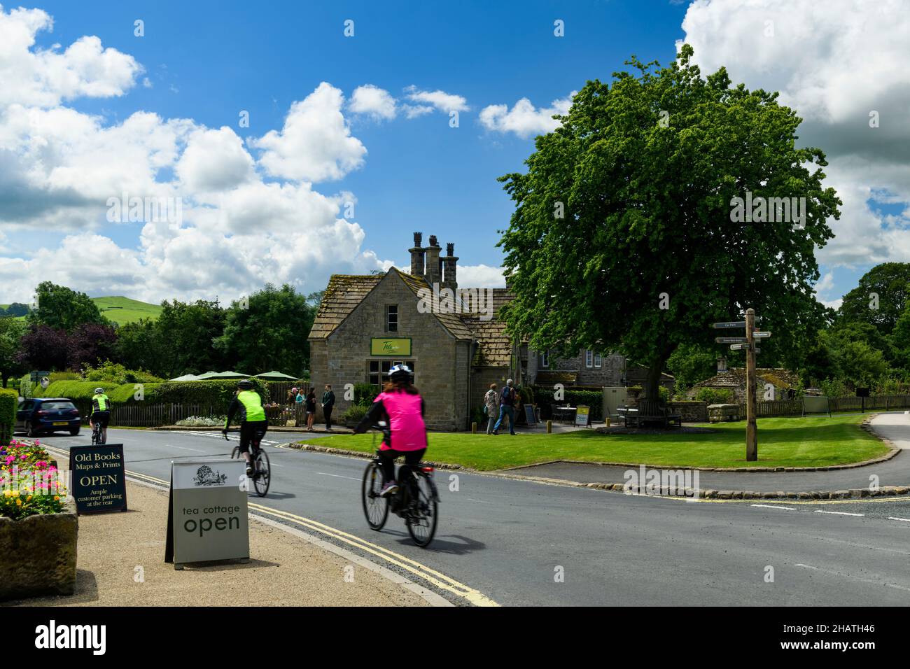 3 cyclistes et marcheurs passant devant un charmant cottage salon de thé café dans un pittoresque village rural ensoleillé - B6160 Bolton Abbey, Yorkshire Dales, Angleterre Royaume-Uni. Banque D'Images