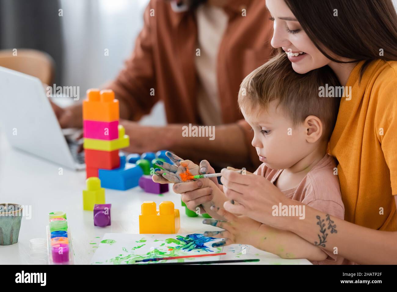 Bonne maman peinture sur la main de fils près des blocs de construction et mari utilisant l'ordinateur portable à la maison Banque D'Images