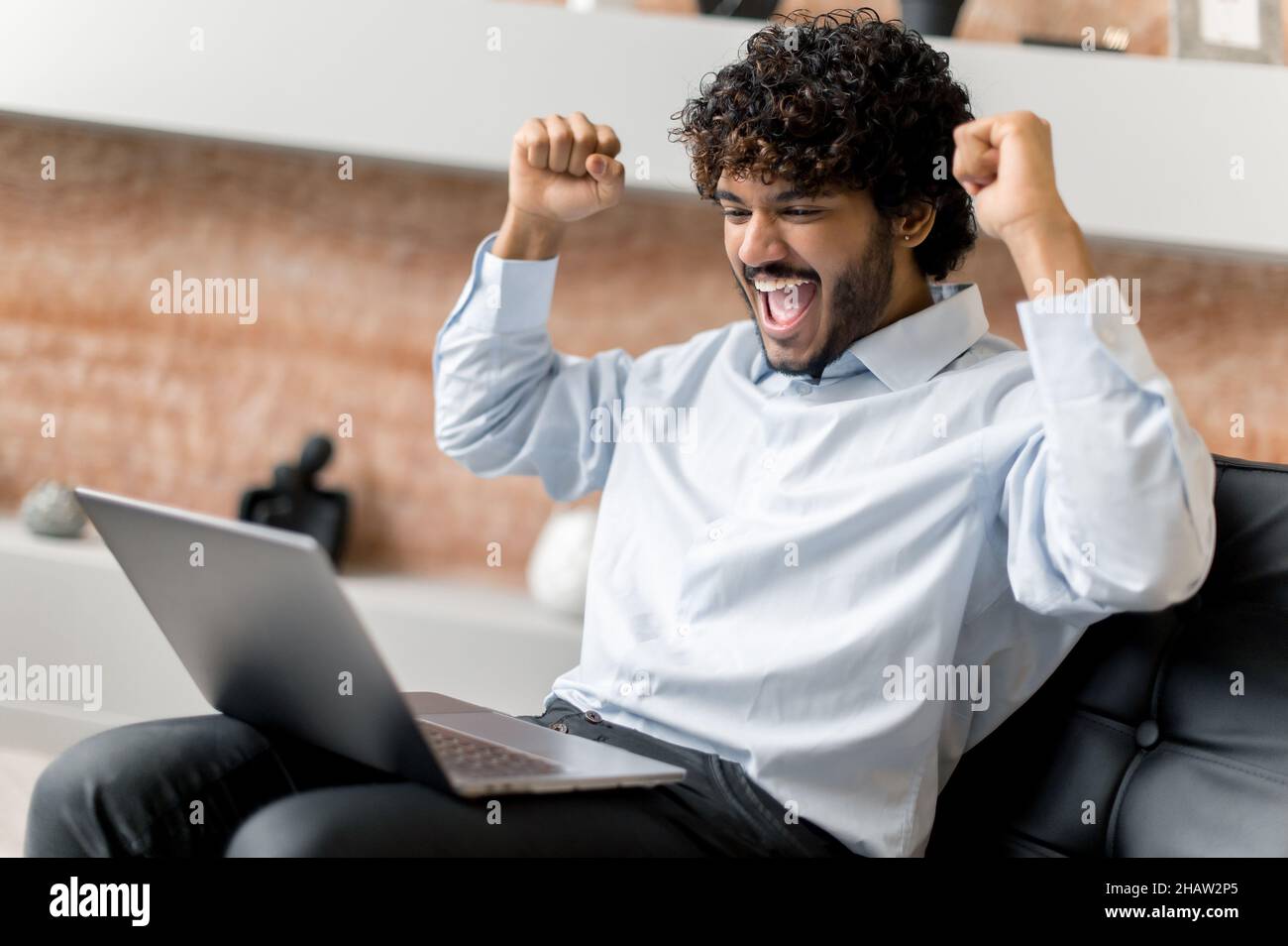 Jeune adulte réussi heureux Indien ou arabe homme d'affaires, courtier ou gestionnaire dans des vêtements formels, assis sur un fauteuil, a eu de bonnes nouvelles, se réjouir de la victoire, bonne affaire,gros profit, gestuelle avec les poings Banque D'Images