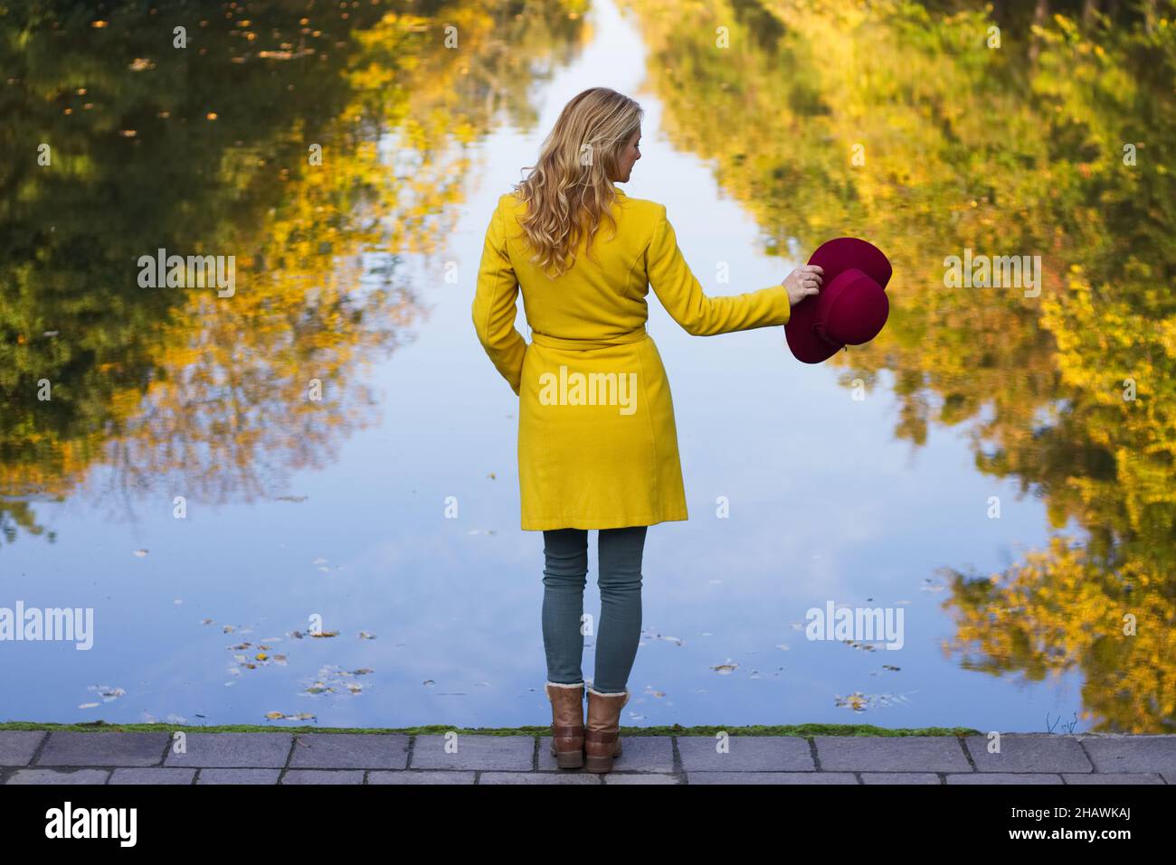 Femme à la mode se tenant au-dessus du lac dans le parc et tient un chapeau rouge.Réflexion des arbres d'automne dans l'eau.Femme tendance portant un manteau de laine. Banque D'Images