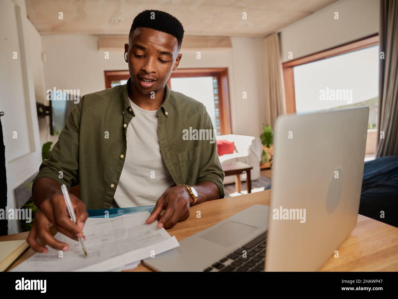 Jeune adulte, homme noir, avec écouteurs, prenant des notes lors d'une réunion en ligne.S'asseoir au comptoir de la cuisine à l'aide d'un ordinateur portable. Banque D'Images