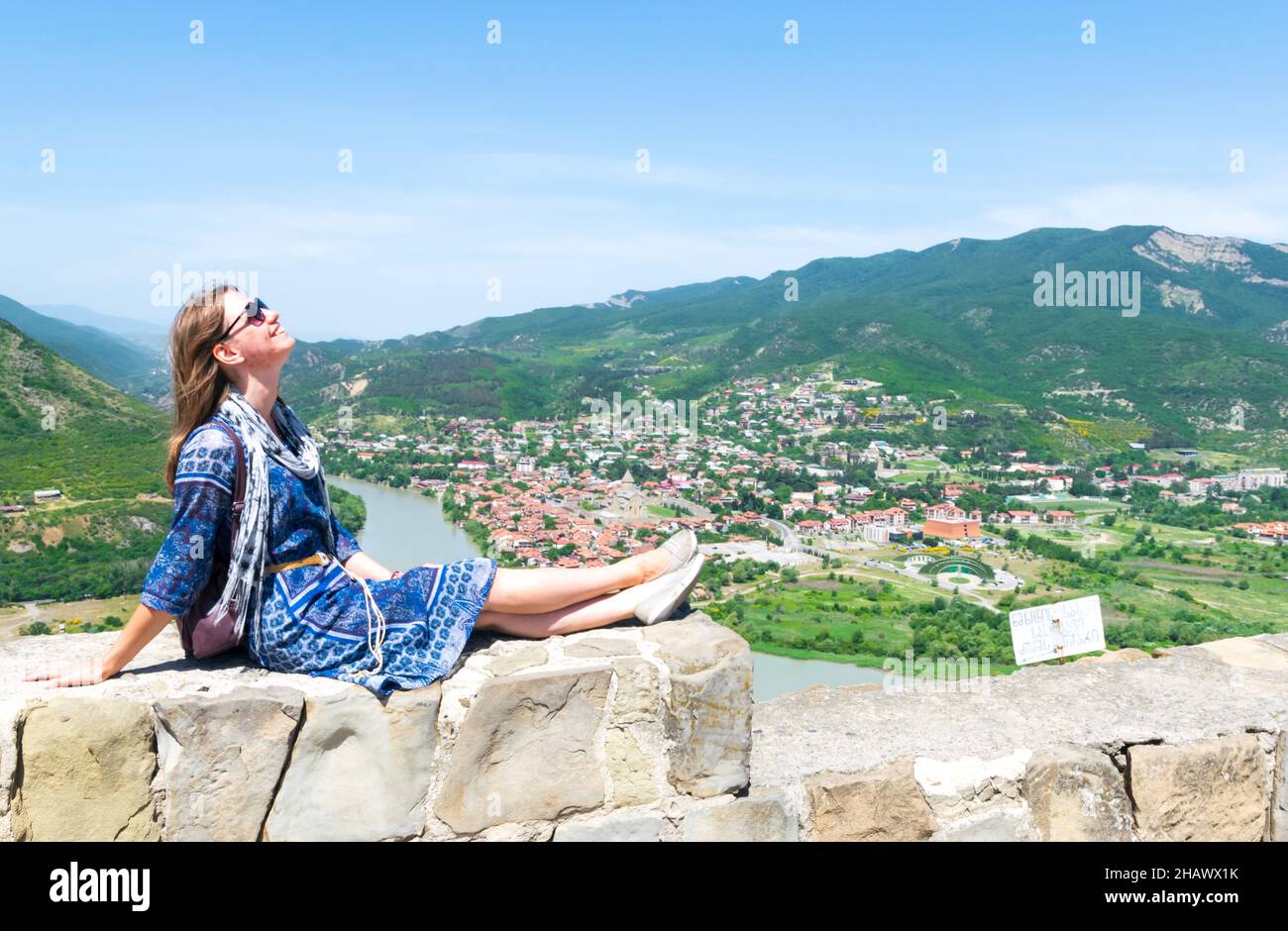 Jeune femme caucasienne touriste en robe se trouve sur le mur de brique pose pour la photo de voyage avec le fond de la ville de Mtskheta Banque D'Images