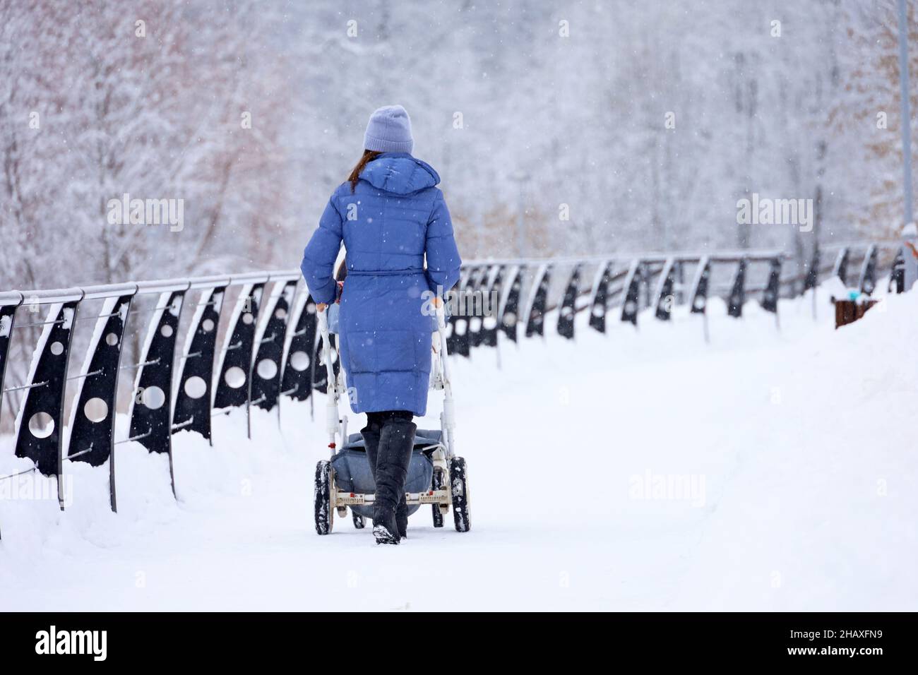 Fille en manteau bleu et bottes marchant avec une poussette de bébé dans le parc d'hiver.Météo neige, concept de maternité, mère célibataire avec pram Banque D'Images