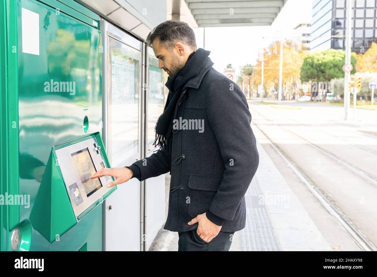 Vue latérale d'un homme sérieux en manteau noir debout avec la main dans la poche et l'achat de billet à la gare dans la ville en journée Banque D'Images