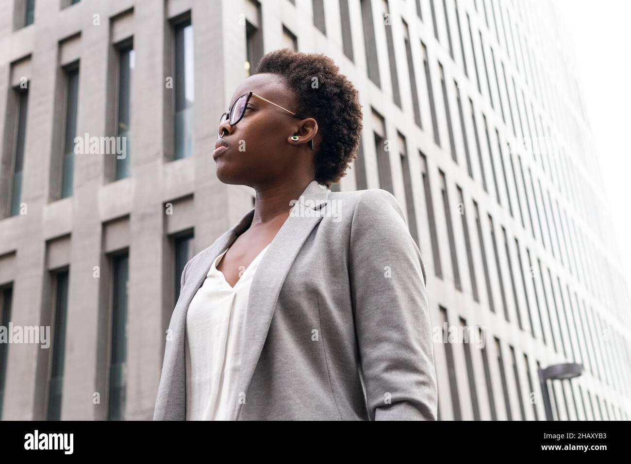 Faible angle de jeune femme entrepreneur en veste élégante et lunettes de vue debout près des gratte-ciels modernes de la ville Banque D'Images