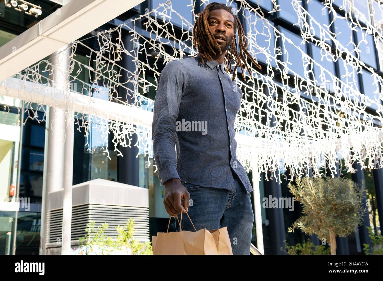 Faible angle d'homme noir confiant avec des dreadlocks et dans des vêtements en denim avec des sacs en papier près du centre commercial avec des guirlandes Banque D'Images