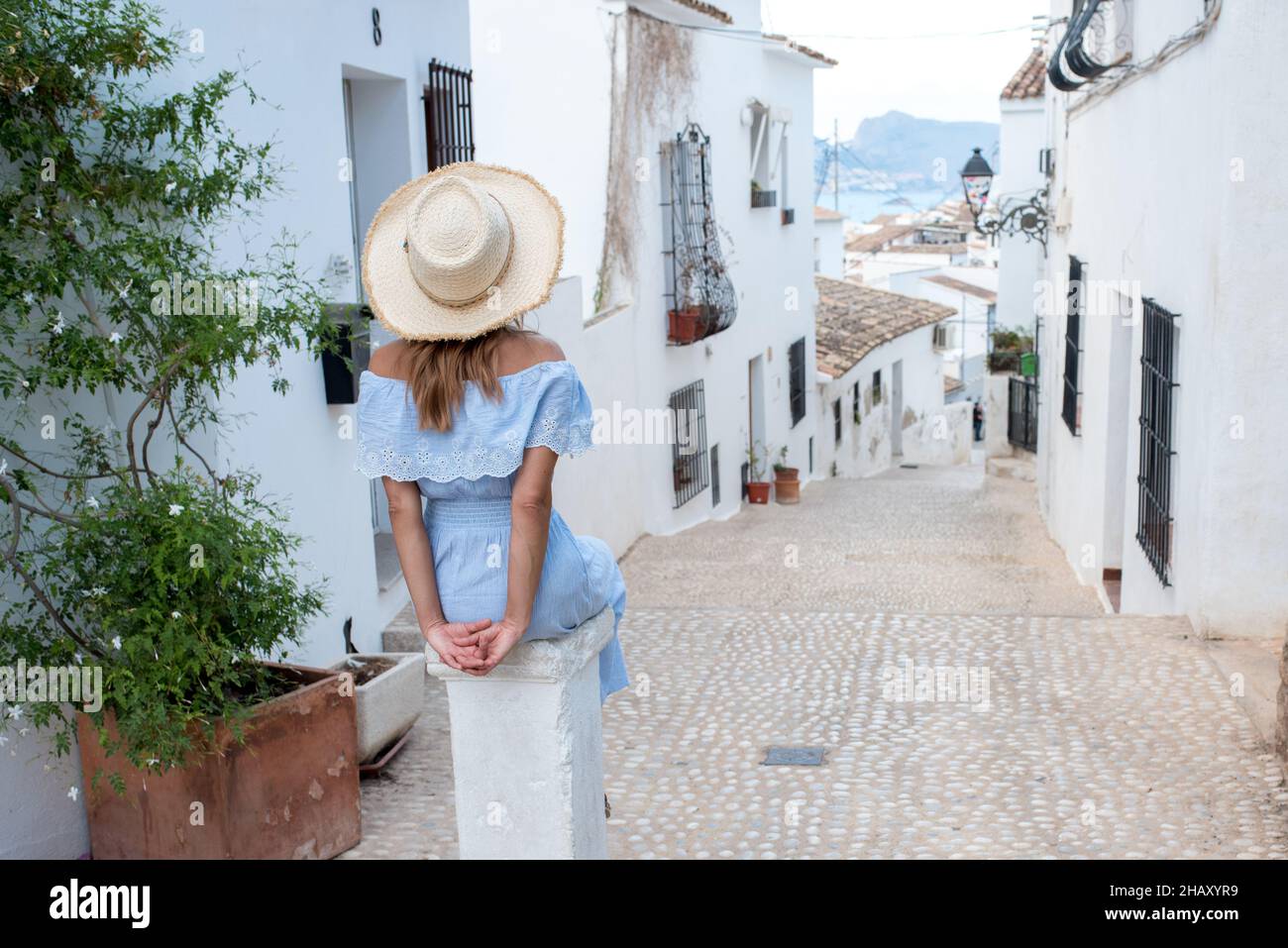 Vue arrière d'une touriste féminine méconnaissable en tenue élégante avec un chapeau assis sur la pierre près des maisons aux intempéries blanches d'Altea pendant le voyage en Espagne Banque D'Images