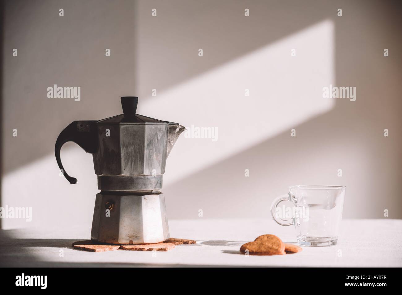 Pot de moka en métal placé sur une table blanche près d'une tasse de verre et de biscuits en forme de coeur le jour ensoleillé Banque D'Images