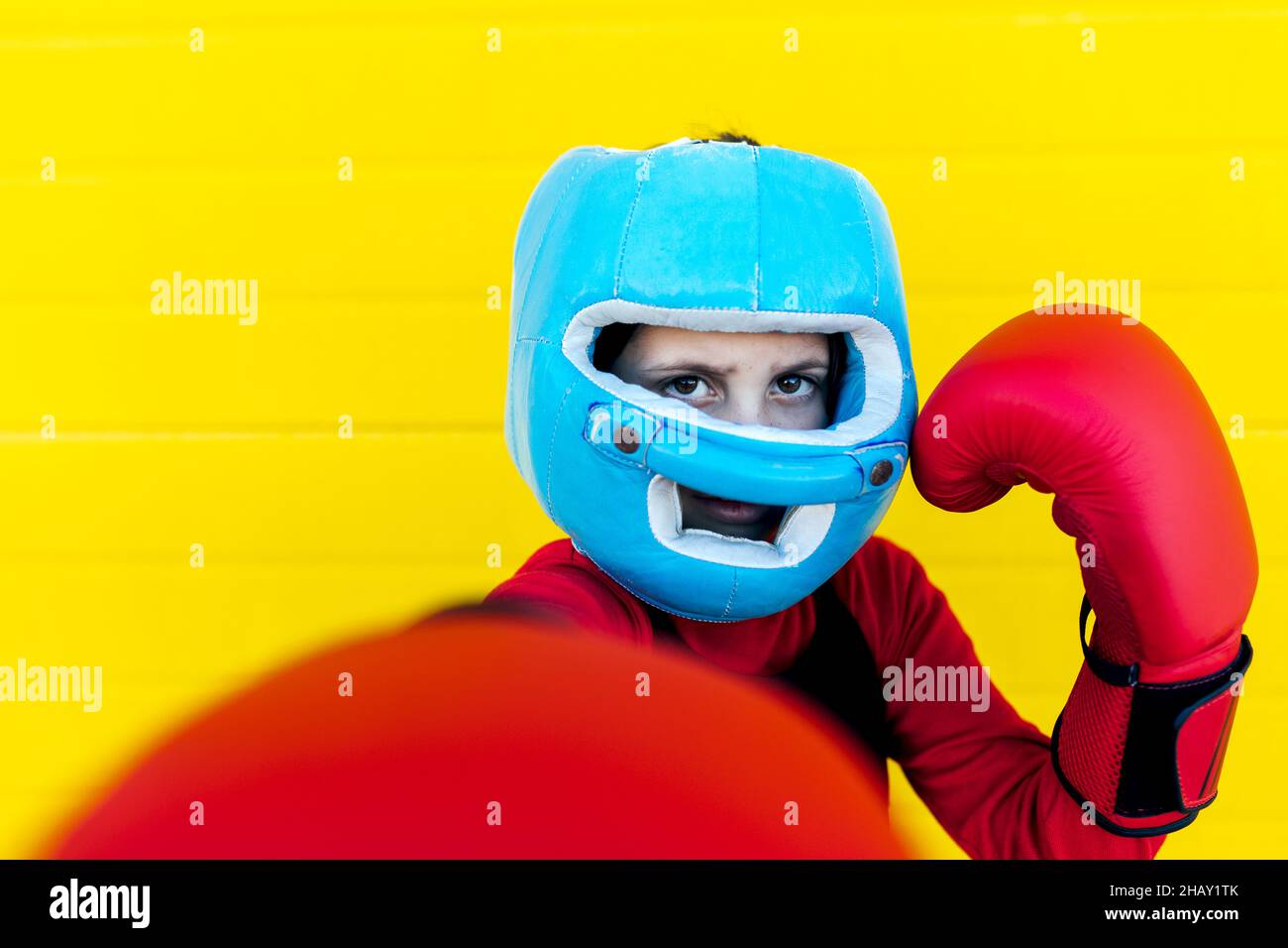 Une fille forte dans un casque de protection levant les bras dans des gants de boxe pour montrer la force près du mur jaune et regarder la caméra Banque D'Images