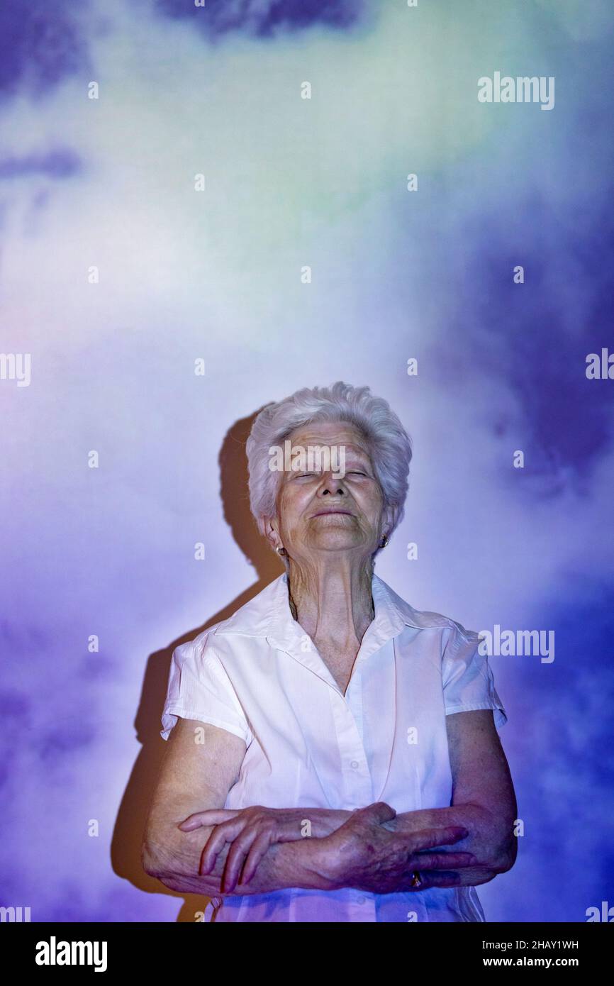 Femme âgée calme aux cheveux gris et aux yeux fermés debout avec des bras croisés en projection de nuages blancs du ciel après la mort Banque D'Images