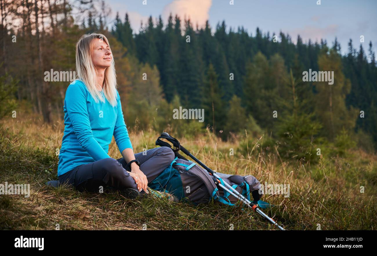 Femme assise sur la pelouse et se reposant pendant la marche avec des bâtons et un sac à dos, regardant avec un sourire détendu et insouciant, respirant de l'air frais.Concept de voyage, randonnée. Banque D'Images