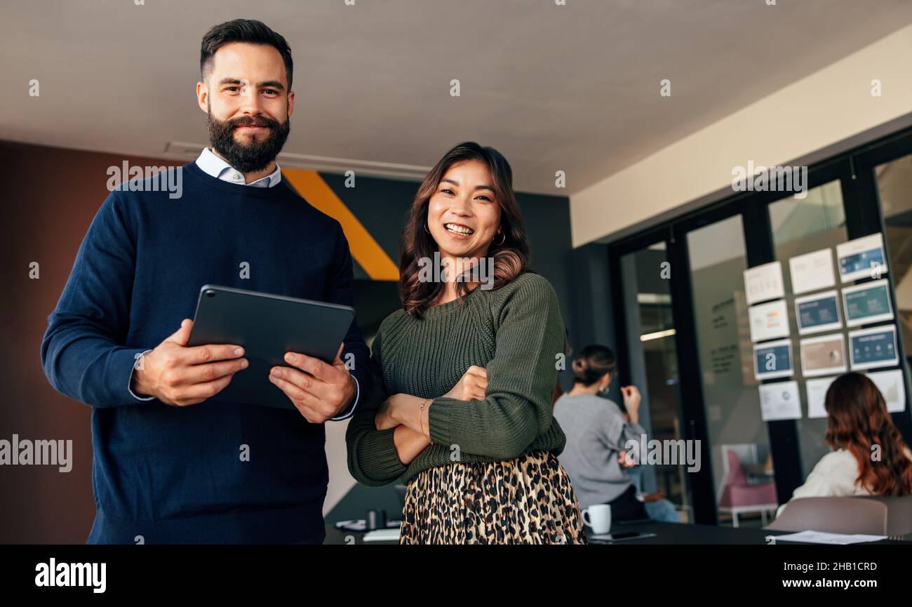 Des collègues d'affaires souriants souriant à l'appareil photo.Deux jeunes hommes d'affaires debout dans une salle de réunion avec leurs collègues en arrière-plan.Divers Banque D'Images