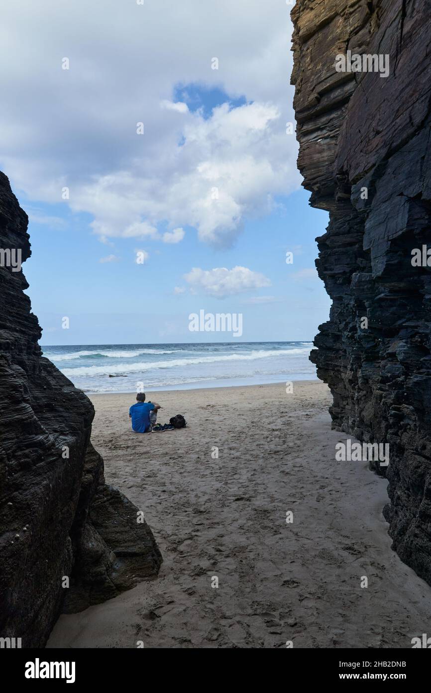 Homme à l'extérieur, par une formation rocheuse, avec ses chaussures de sport et son sac à dos déposés à ses côtés, il contemple la mer assis sur le sable de la Banque D'Images