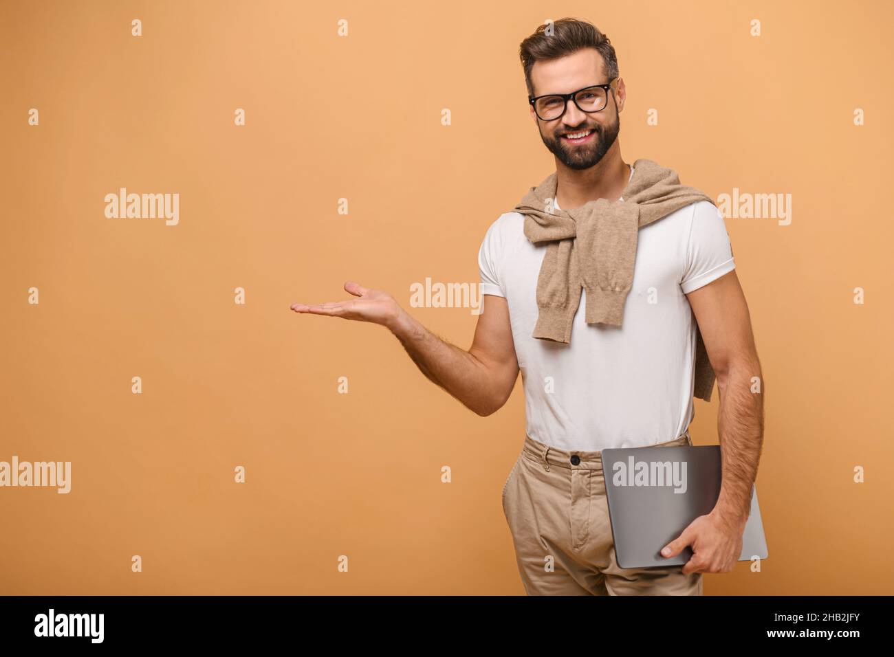 Homme positif indépendant avec la barbe tenant un ordinateur portable et montrant à l'endroit vide avec la main, satisfait du télétravail, aime son travail.Studio d'intérieur isolé sur fond orange Banque D'Images