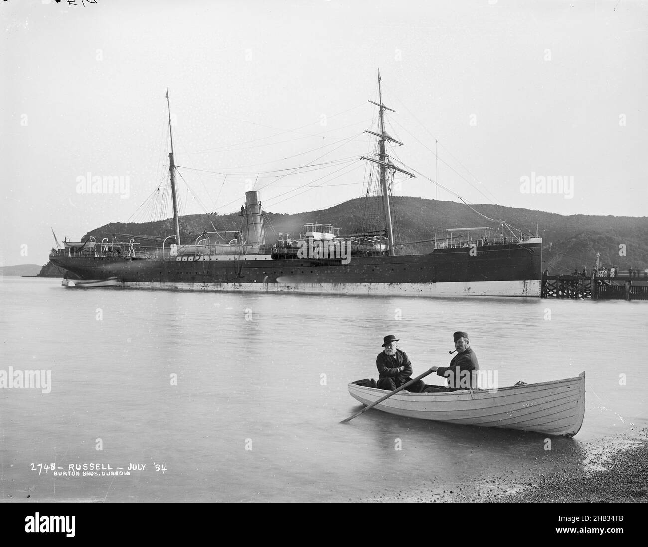 Russell - juillet 84, Burton Brothers studio, studio de photographie, juillet 1884, Nouvelle-Zélande,Photographie en noir et blanc, Foreshore, avec deux hommes sur un bateau à rames, un aviron avec pipe dans la bouche.Grand navire avec deux mâts et un entonnoir, un bateau est amarré à sa poupe, est amarré à un quai et une colline derrière est en arrière-plan.Le bateau est le 'Wairararapa Banque D'Images