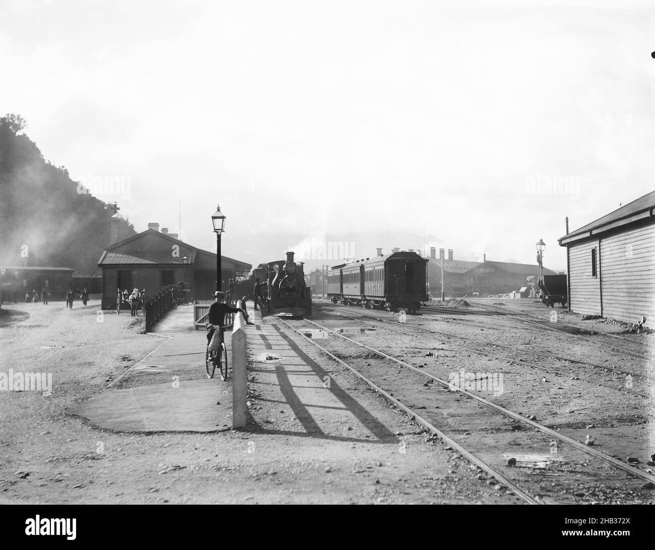 [Gare de Greymouth], studio Muir & Moodie, studio de photographie, Nouvelle-Zélande, gare de Greymouth prise de l'extrémité Hokitika de la plate-forme principale.À gauche, un train est récemment arrivé à la plate-forme Riverside et les passagers débarqués traversent l'intersection de la rue Mackay et du quai de Mawhera.Sur la droite, en arrière-plan derrière les calèches, se trouvent les anciennes hangars de moteurs de Greymouth qui ont été démolis vers 1929.Le hangar, au premier plan droit, a également été démoli avant 1969 Banque D'Images