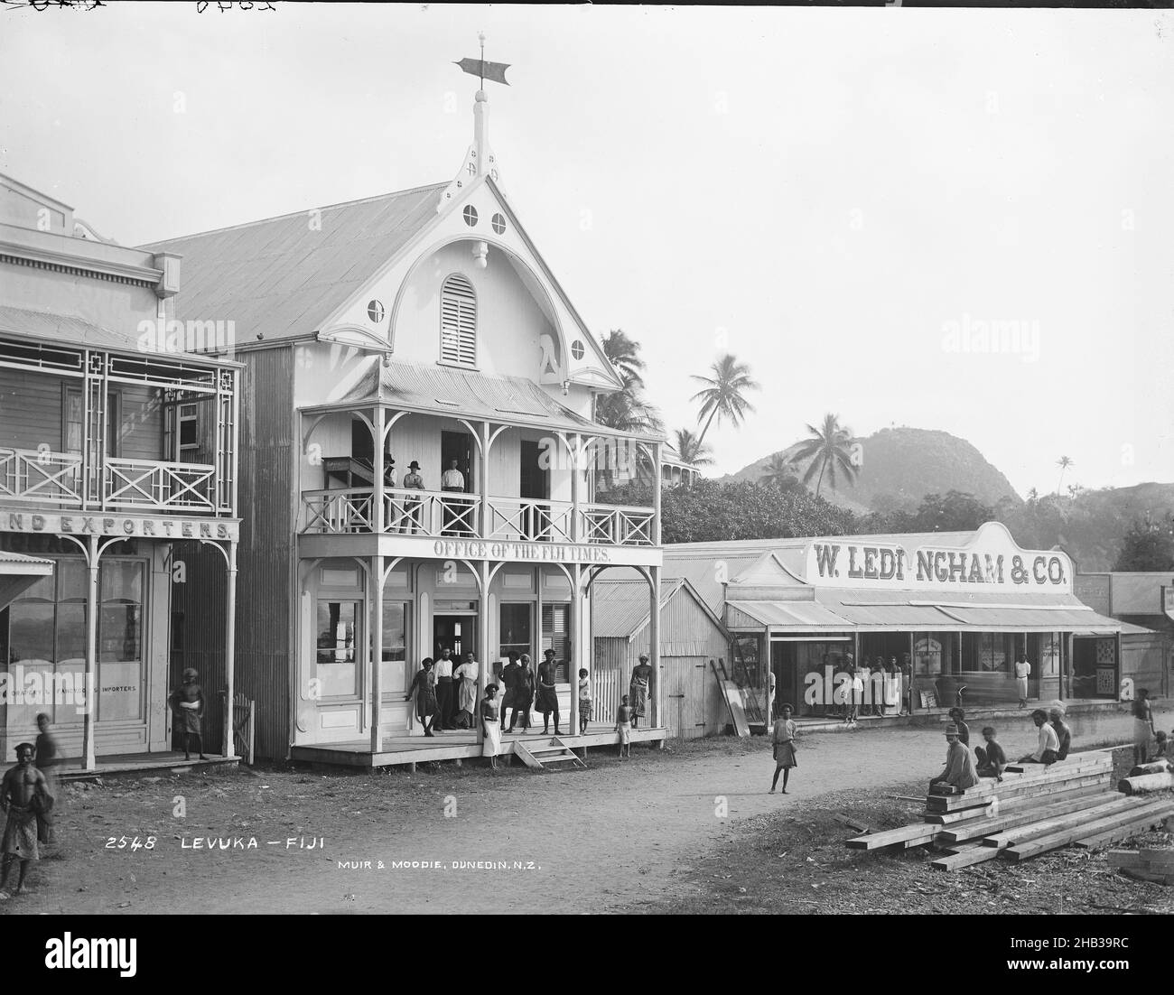 Levuka, Fidji, Burton Brothers studio, studio de photographie, juillet 1884,Nouvelle-Zélande, photographie en noir et blanc, route avec boutiques de la ville.Deux bâtiments de 2 étages avec des vérandahs et deux autres bâtiments, en face est une pile de bois sur lequel les jeunes enfants et un adulte sont assis.Le bâtiment principal (2 étages) est doté d'un panneau Office of the Fiji Times et, à côté, d'un seul étage avec panneau W. Ledingham & Co.Le fond est une colline avec des cocotiers Banque D'Images