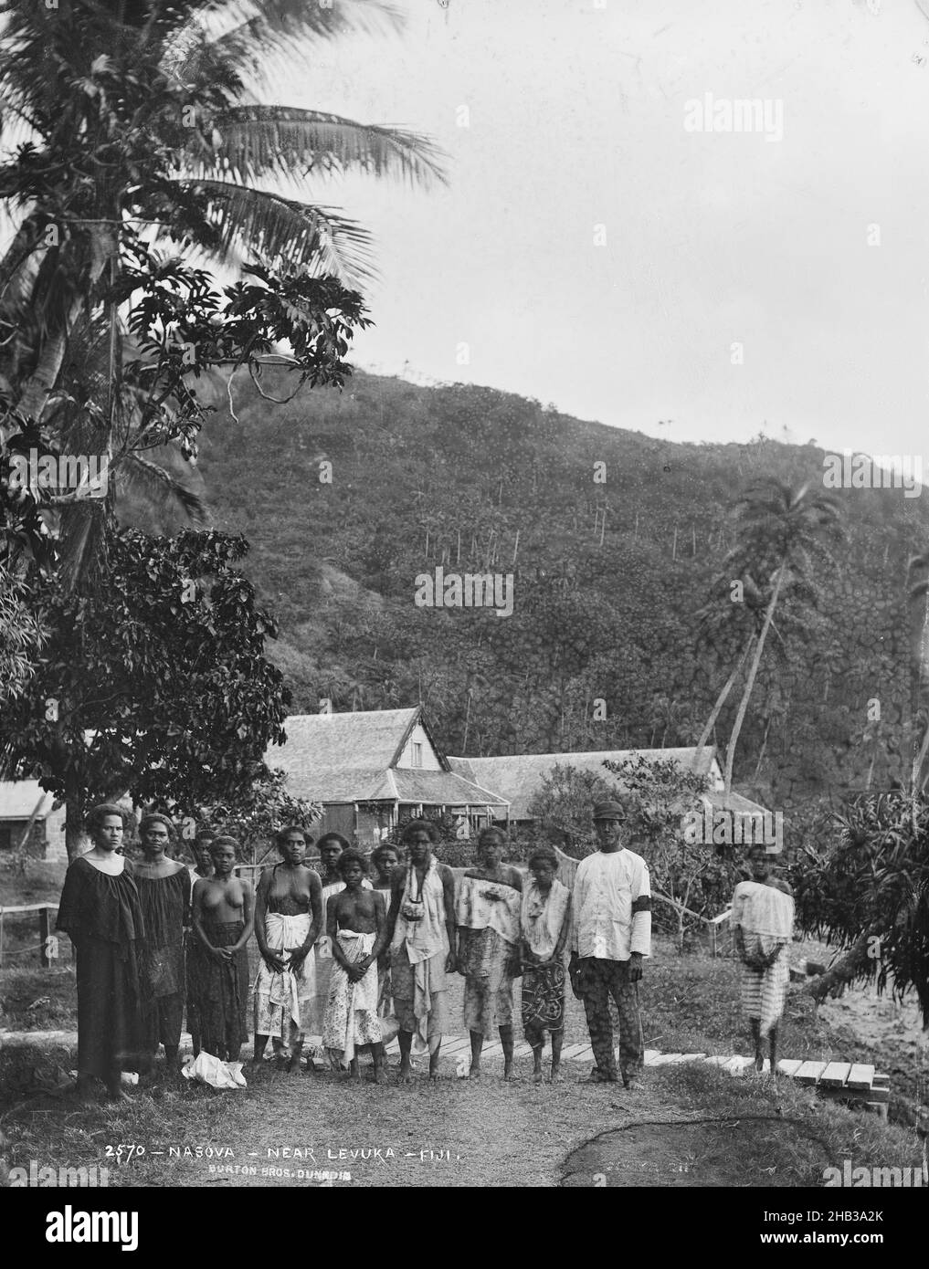 Nasova, près de Levuka, Fidji, Burton Brothers studio, studio de photographie,14 juillet 1884, Nouvelle-Zélande, photographie en noir et blanc, 11 femmes debout dans deux rangées, un homme en uniforme en fin de rangée (à droite), puis un écart avec une femme debout seule.Tous sont fidjiens.Une route s'étend de l'avant-plan à l'arrière-plan des gens.Une clôture et une rangée de maisons coloniales sont visibles.Les cocotiers s'encourent jusqu'à une haute colline qui s'élève fortement en arrière-plan Banque D'Images