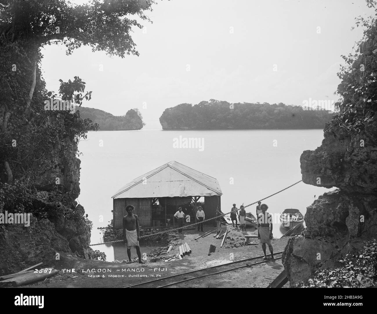 La lagune, Mango [Mago], Fidji, Burton Brothers studio, studio de photographie,Juillet 1884, Dunedin, photographie en noir et blanc, hangar de bateau sur le rivage du lagon, avec ligne de chemin de fer devant.Deux masses terrestres peuvent être vues en arrière-plan.Deux longs bateaux sont à côté de la Boathouse, et trois hommes sont directement devant et trois à droite.Deux hommes fidjiens se tiennent près du centre en amont et près du premier plan.Au premier plan de chaque côté se trouvent des arbres et une face rocheuse à droite Banque D'Images