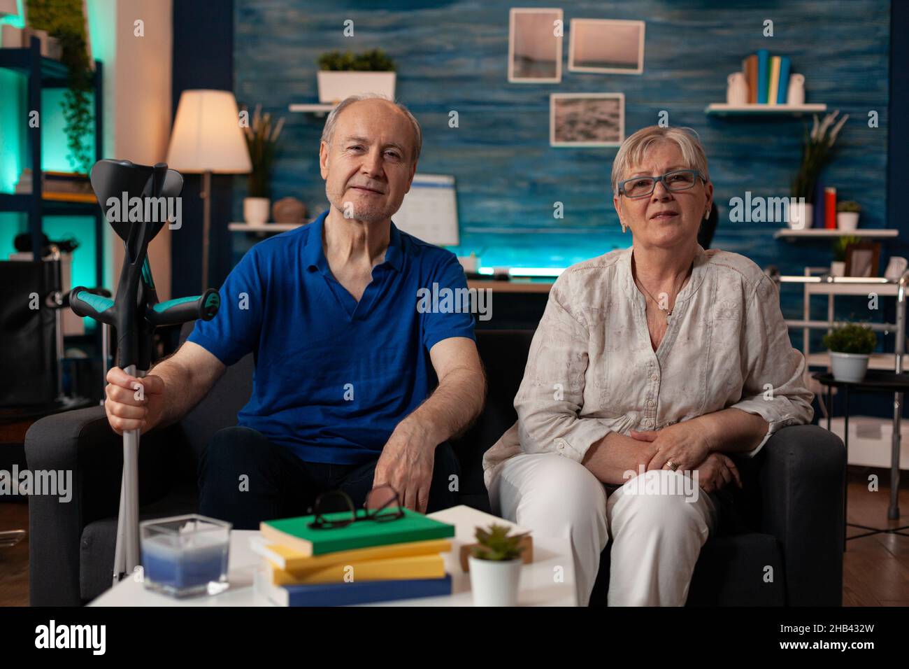 POV de vieux couple marié assis sur le canapé de salon ensemble tout en regardant la caméra.Les retraités caucasiens se détendent à la maison sur un canapé et profitent de leur retraite.Homme senior tenant des béquilles Banque D'Images