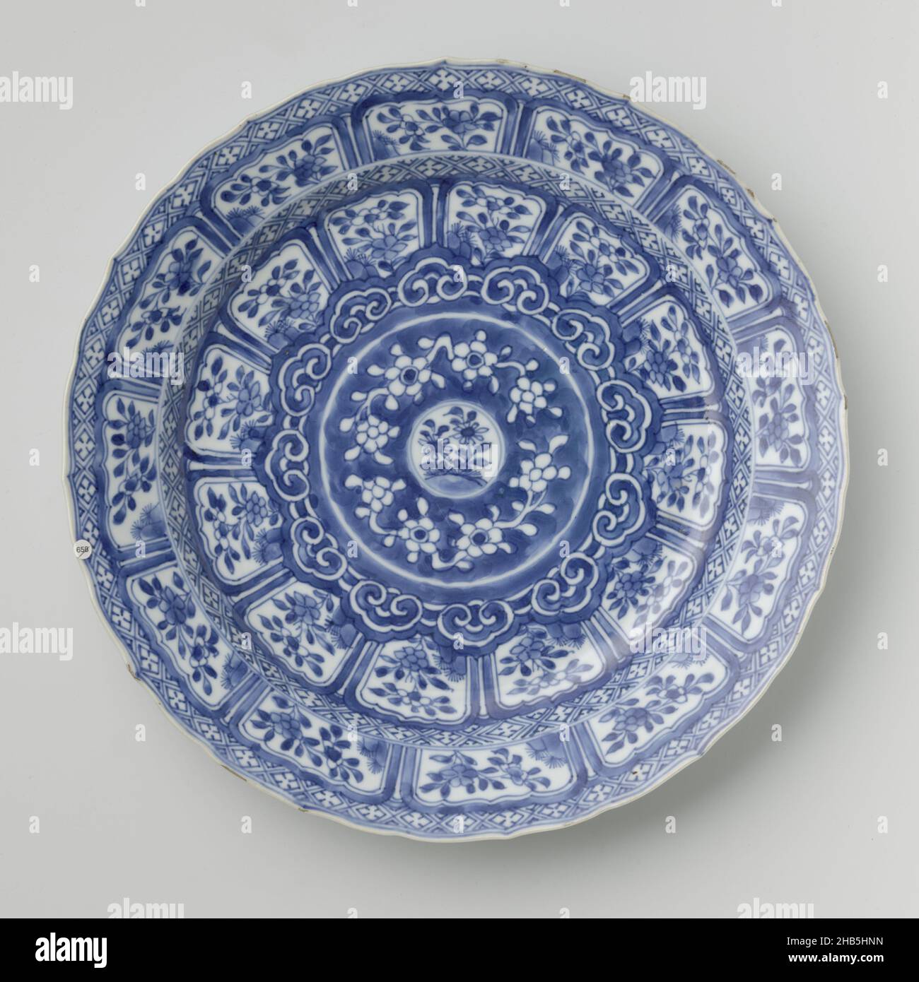 Plat au médaillon entouré de bordures ornementales, plat en porcelaine avec paroi surélevée et rebord incliné, festonné, peint en bleu sous-glaçure.Sur l'appartement un petit médaillon avec une plante à fleurs entourée de plusieurs bordures décoratives avec: Tendrils de fleur, motif ruyi, branche de fleur avec des rochers et servetwork; l'inverse avec deux branches de fleur; sur le fond un double cercle.Bleu et blanc., anonyme, Chine, c.1700 - c.1724, porcelaine, glaçure, peinture, vitrification,hauteur 4,1 cm, diamètre 28,1 cm, diamètre 17 cm Banque D'Images