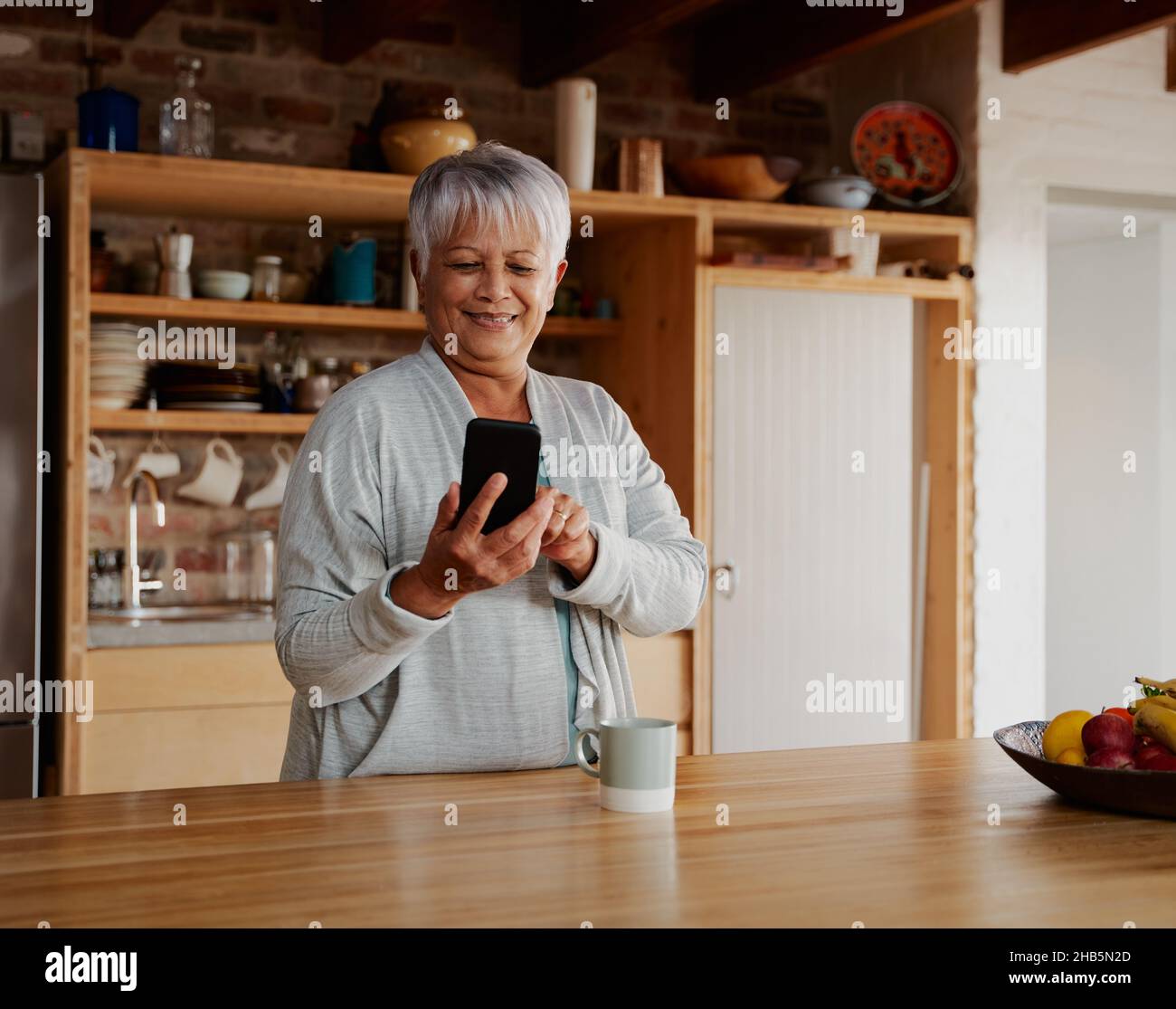 Orientation portrait d'une femme âgée, multiculturelle et heureuse, défilant sur un smartphone tout en étant debout dans une cuisine moderne. Banque D'Images