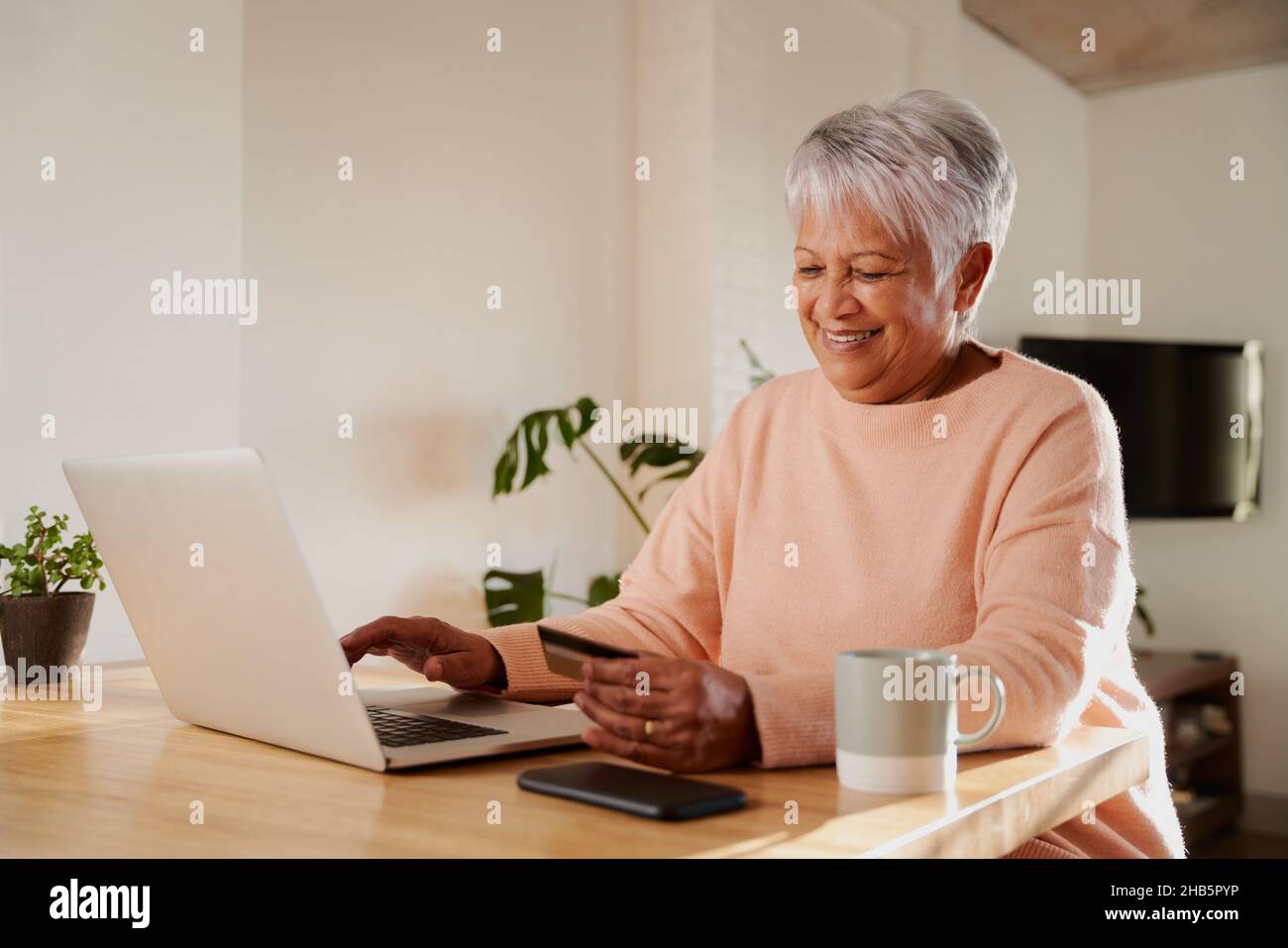 Une femme âgée de plusieurs ethnies se fait un plaisir de payer en ligne sur un ordinateur portable, assise au comptoir de la cuisine. Banque D'Images
