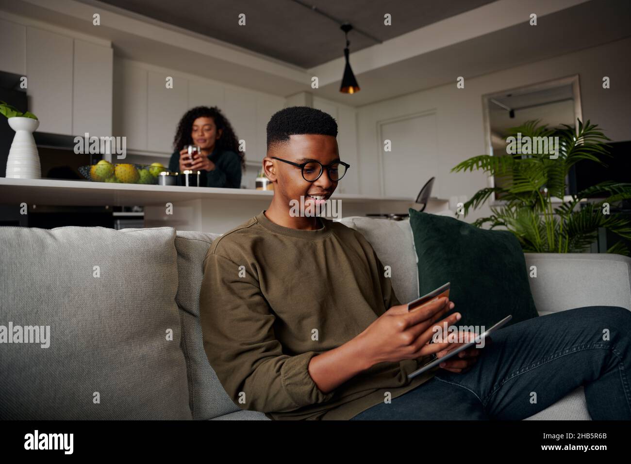 Noir homme faisant l'achat en ligne sur le canapé tandis que la petite amie est dans la cuisine, détendu et heureux Banque D'Images Noir homme faisant l'achat en ligne sur le canapé tandis que la petite amie est dans la cuisine, détendu et heureux Banque D'Images