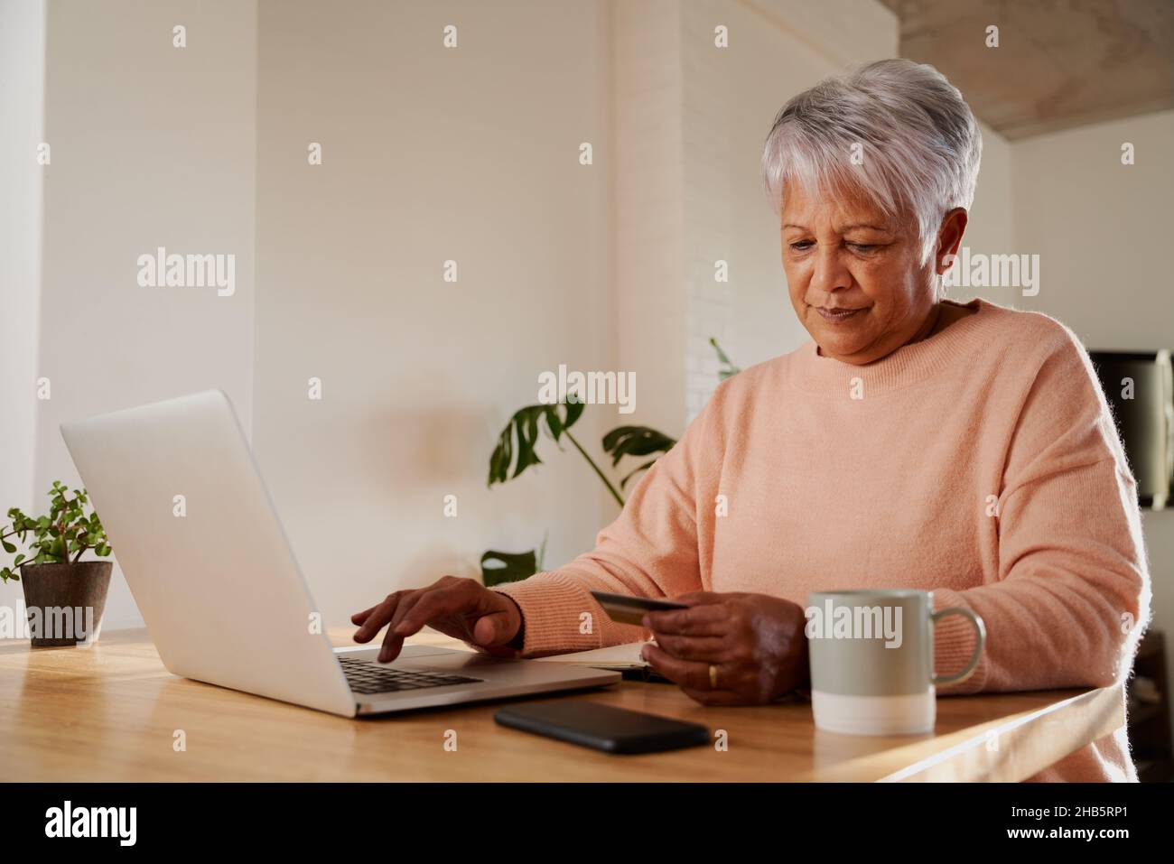 Une femme âgée de plusieurs ethnies se fait un plaisir de taper les coordonnées bancaires pour effectuer un paiement en ligne sur ordinateur portable, au comptoir de la cuisine. Banque D'Images