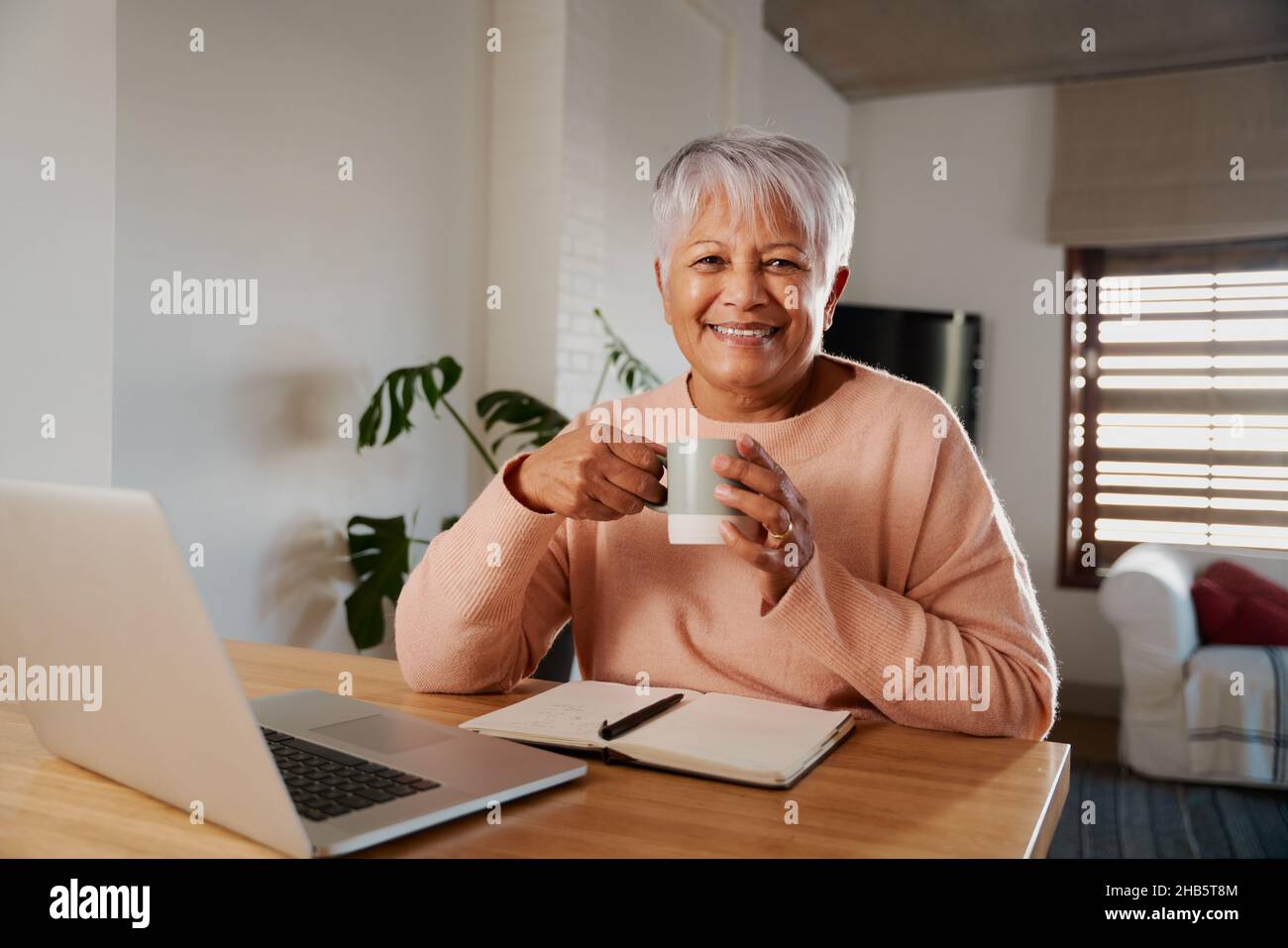 Portrait d'une femme multiethnique âgée souriant à l'appareil photo, tenant une tasse de café du matin dans les mains.Salon avec ordinateur portable au comptoir de la cuisine. Banque D'Images
