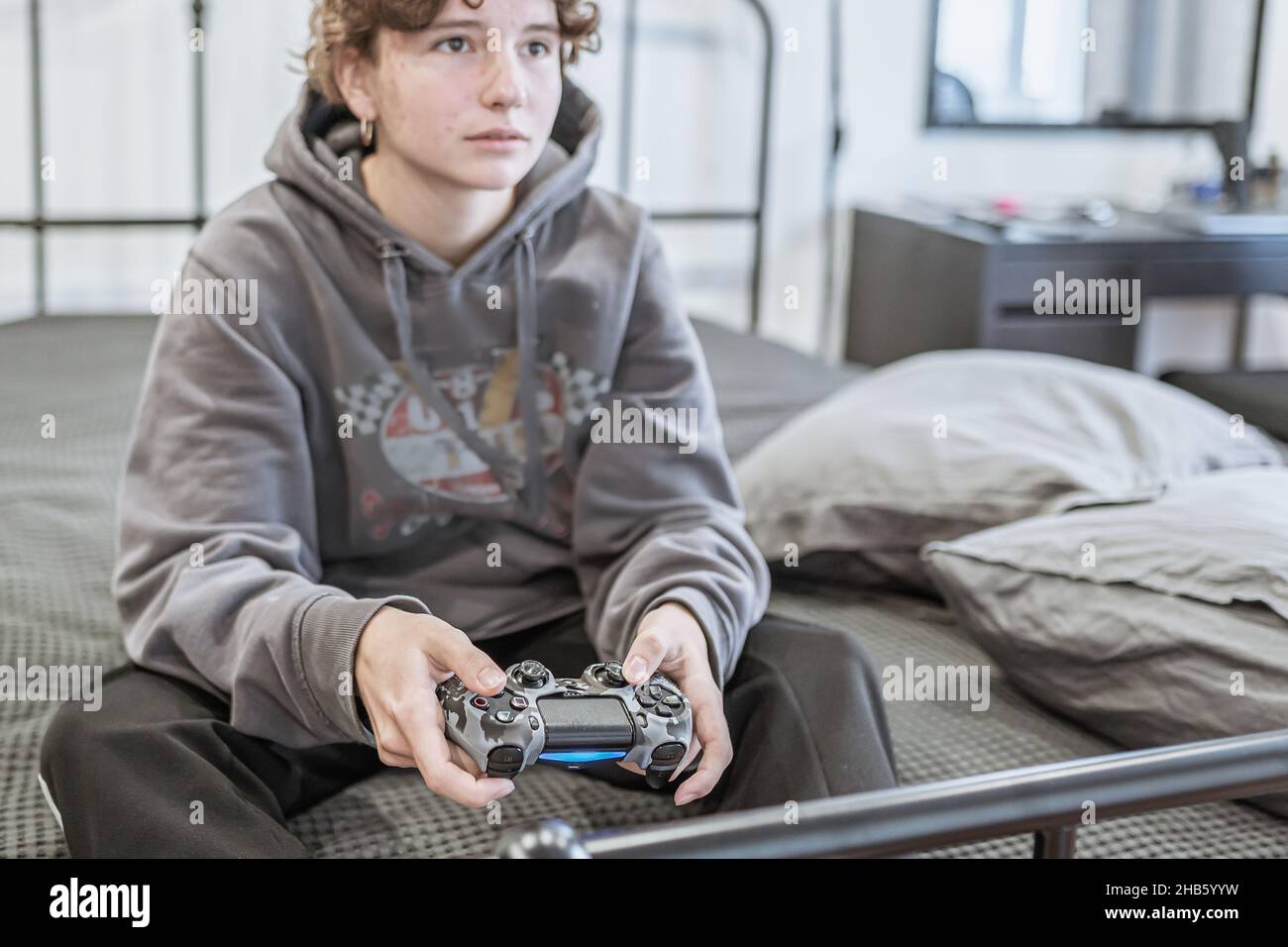 une adolescente aux cheveux bouclés bruns courts, vêtue de vêtements de maison peu habillés, s'assoit sur un lit non fait et joue sur une console d'ordinateur.Passion pour l'ordinateur Banque D'Images