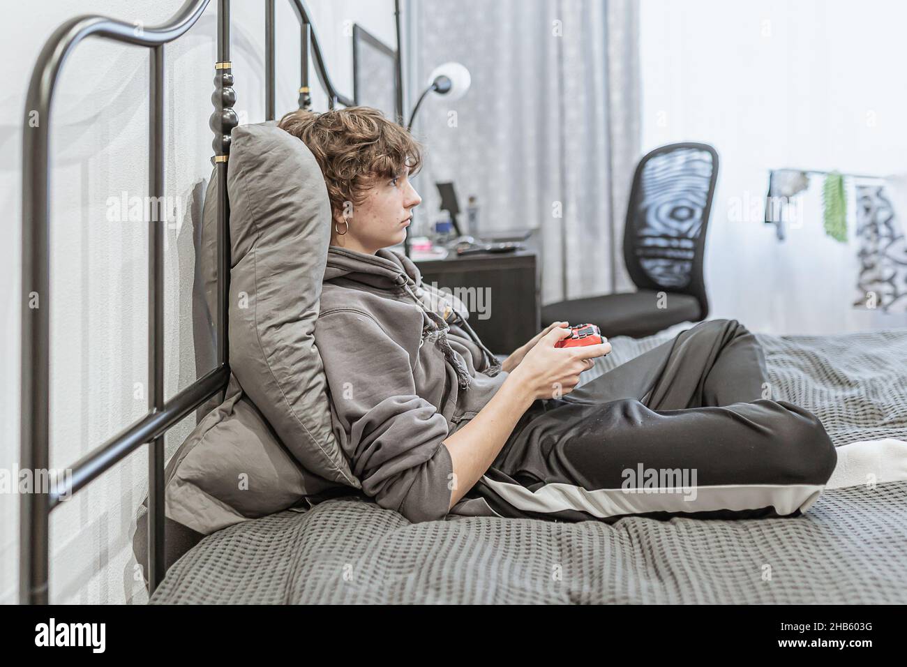 une adolescente aux cheveux bouclés bruns courts, vêtue de vêtements de maison peu habillés, s'assoit sur un lit non fait et joue sur une console d'ordinateur.Passion pour l'ordinateur Banque D'Images