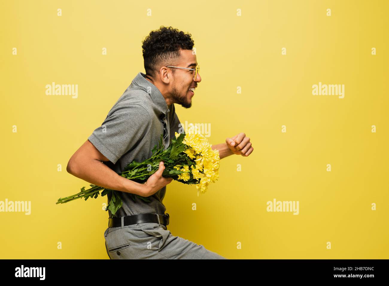 vue latérale de l'homme afro-américain heureux en lunettes de soleil tenant un bouquet de fleurs et marchant sur le jaune Banque D'Images