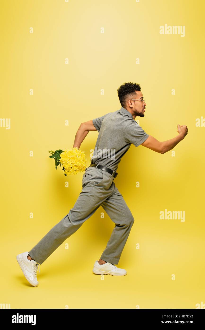 vue latérale de l'homme afro-américain en lunettes de soleil tenant un bouquet de fleurs et courant sur le jaune Banque D'Images
