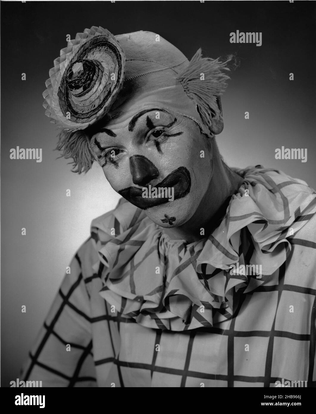 Clown dans une chemise à damier avec col à volants et un petit chapeau de paille posant avec la tête vers le bas et regardant la caméra.Vers 1955 Banque D'Images