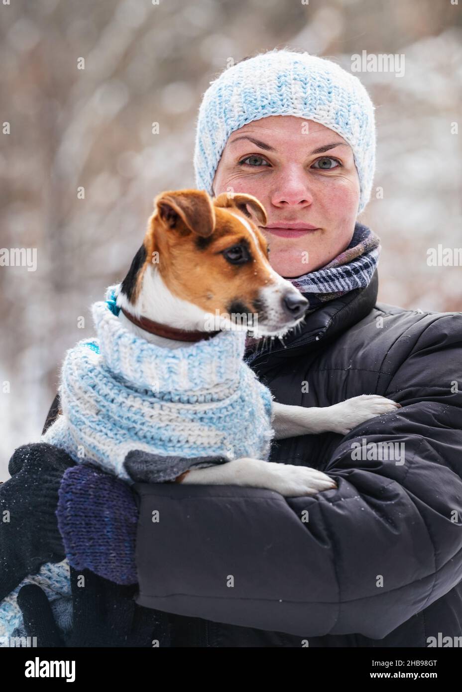 Jeune femme en blouson d'hiver tenant son chien terrier Jack Russell portant des vêtements chauds d'hiver sur les mains, neige floue couvert arbres arrière-plan Banque D'Images