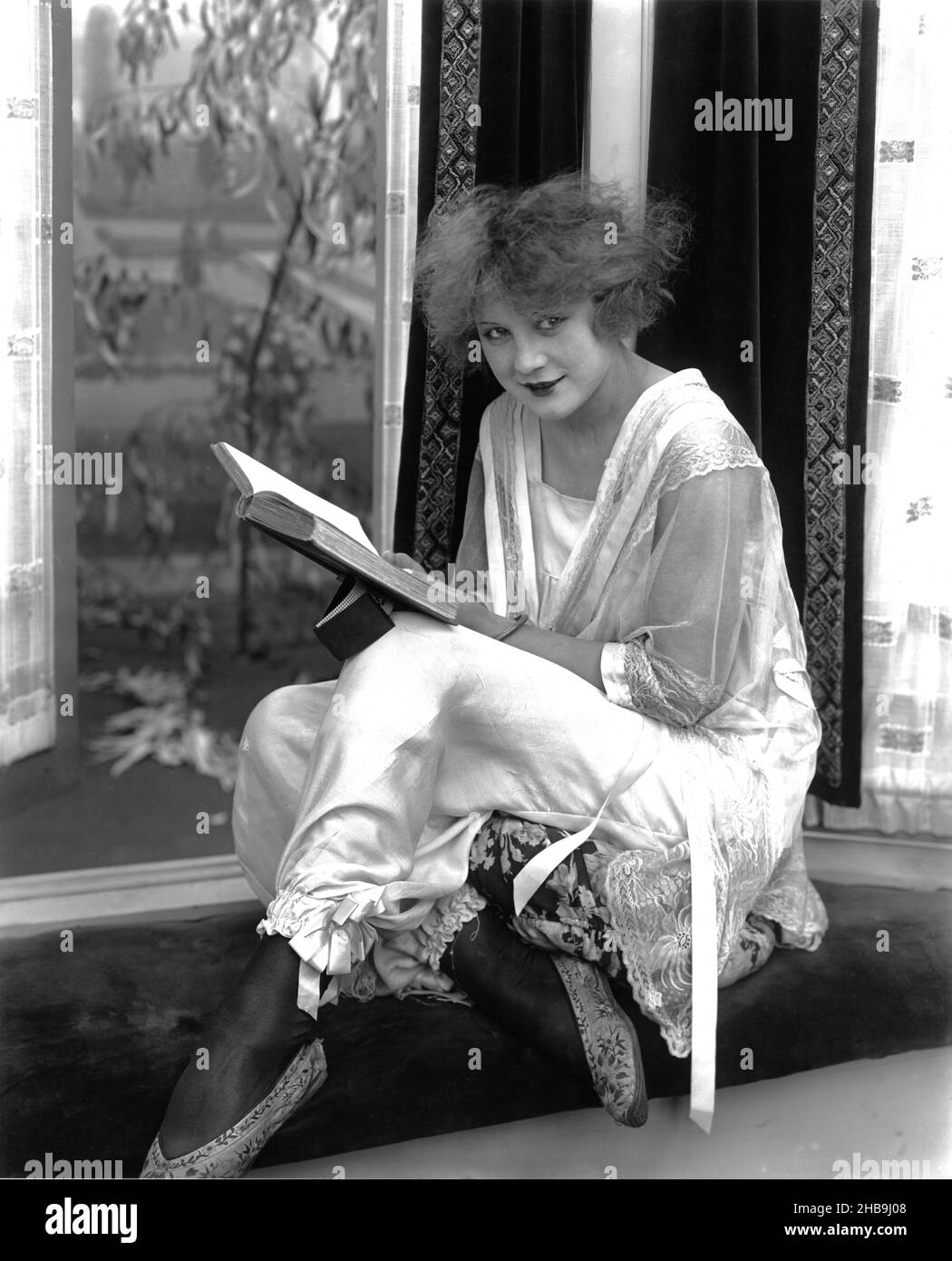 L'actrice Marvel Rae (11/9/1901 - 6/17/1937) dans une personne négligée assise devant une fenêtre faisant une pause de la lecture d'un livre à poser pour la caméra.Vers 1935 Banque D'Images