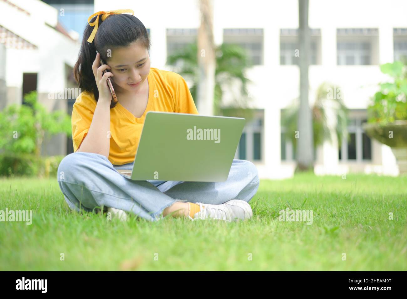 Jeune femme assise sur la pelouse parlant au téléphone et à l'aide d'un ordinateur portable, portant une chemise jaune et un Jean, visage souriant, ordinateur portable sur les jambes, travail de hom Banque D'Images