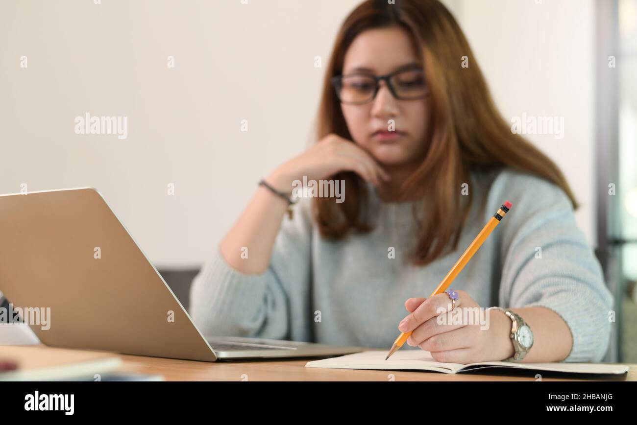 Une adolescente portant des lunettes utilise un crayon pour écrire sur un bloc-notes avec un ordinateur portable sur le bureau, une étudiante d'université prenant des notes en ligne d'un tour Banque D'Images