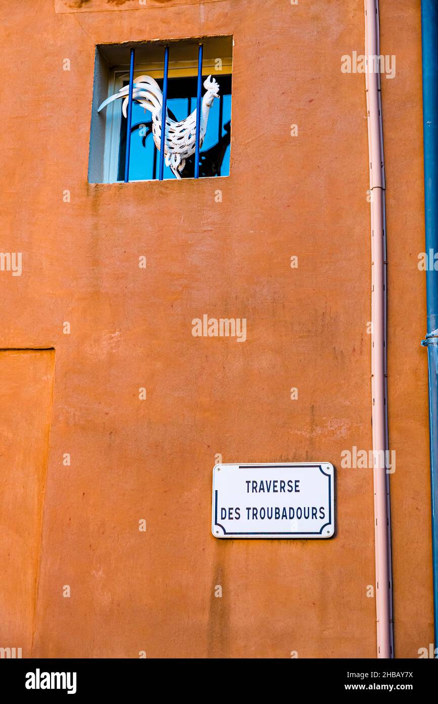 Une grille de fenêtre en fer forgé sur un mur ocre d'une maison à Bormes-les-Mimosas, dans le Var, encadre la silhouette d'un coq ; en dessous, une plaque de rue marque la « Traverse des Troubadours » (Croisement des Troubadours). Banque D'Images